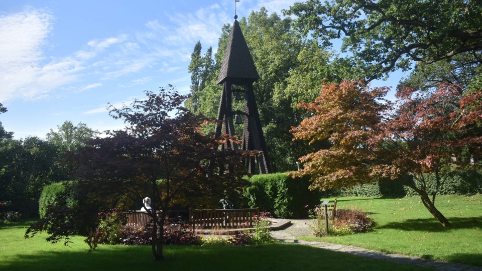 En hög klockstapel i trä bland träd och rabatter.