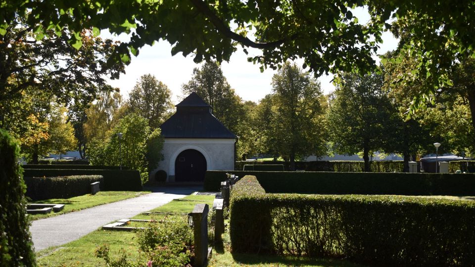 En liten vit kyrkobyggnad på en begravningsplats med gravar och häckar.
