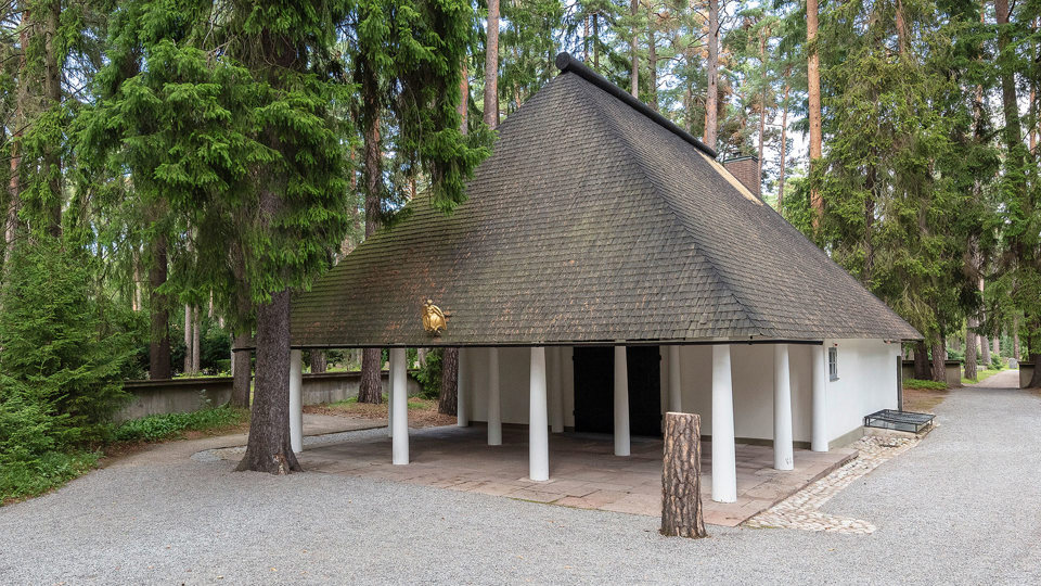 En vit kapellbyggnad med högt spetsigt tak omgivet av höga träd.