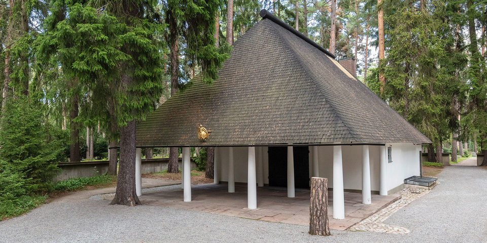 En vit kapellbyggnad med högt spetsigt tak omgivet av höga träd.