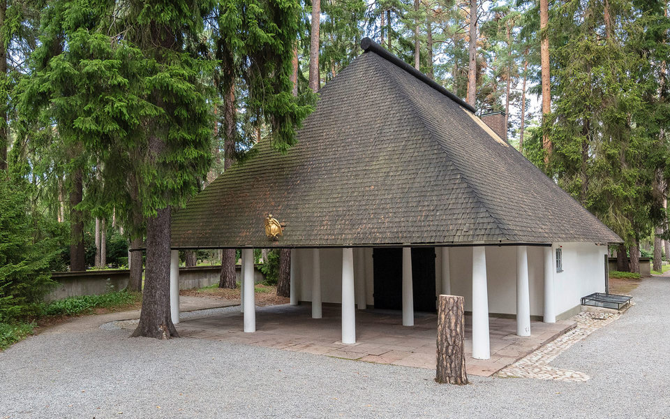 En vit kapellbyggnad med högt spetsigt tak omgivet av höga träd.